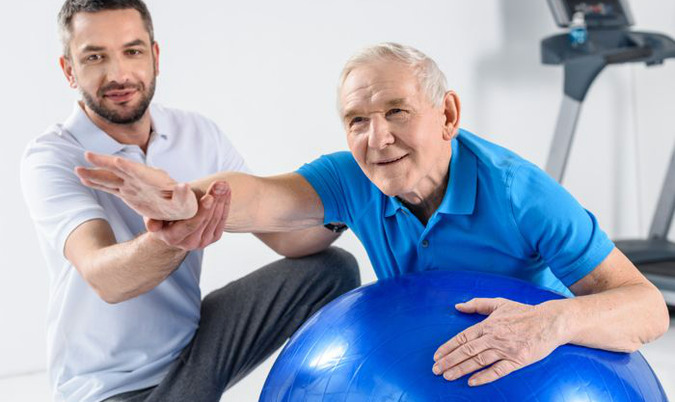 Äldre man får hjälp att hålla balansen på en rund blå boll. 
